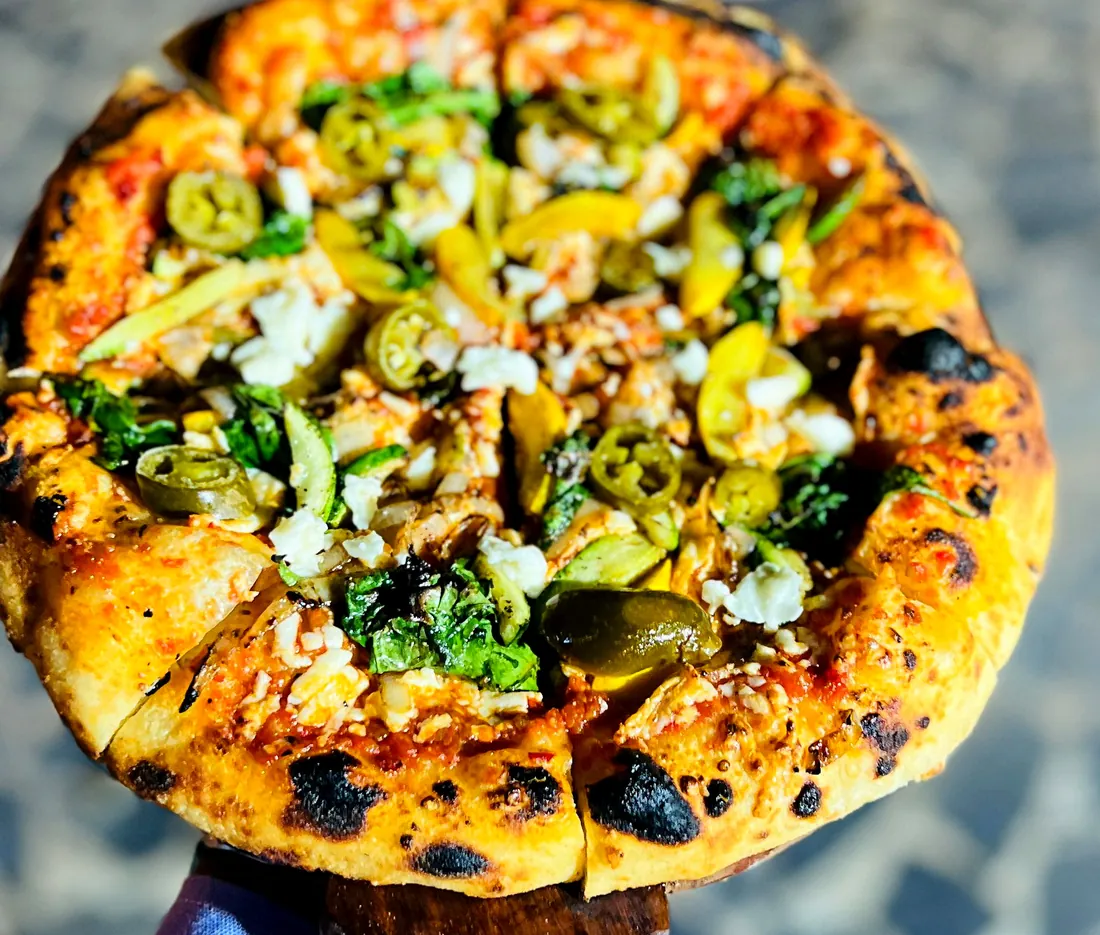 Wood-fired pizza with charred crust, jalapeños and fresh herbs held up to the light